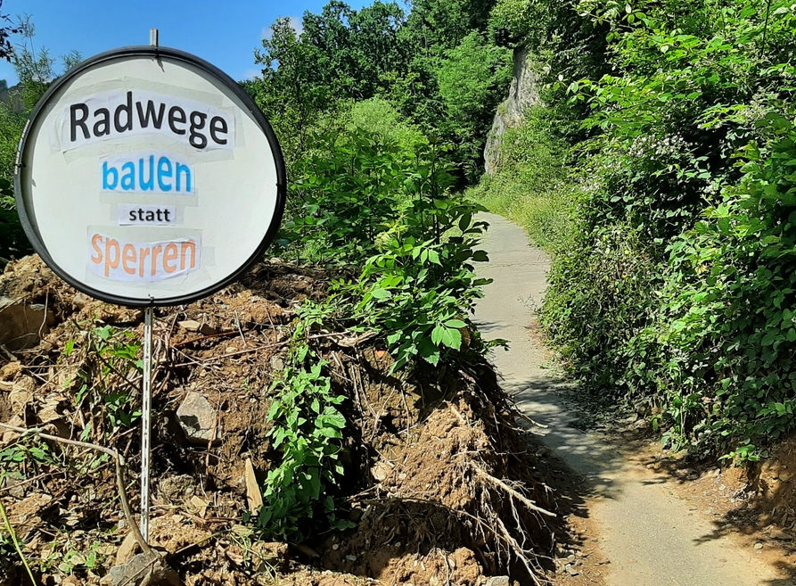 Der Siegtalradweg in der Nähe von Dreisel, welcher mit einer künstlichen Barriere aus Sand, Geröll und Wurzelwerk versperrt wurde. Auf der rechten Seite wurde die Barriere allerdings durchbrochen und ist wieder passierbar, links daneben ein selbstgebasteltes Schild mit der Aufschrift "Radwege bauen statt sperren".