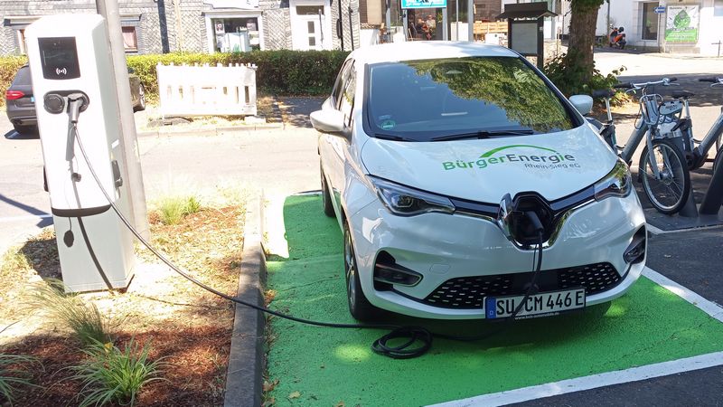 Auf der linken Seite eine Ladesäule an der das Auto rechts daneben angeschlossen ist.