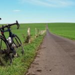 Ein Fahrrad lehnt an einem Zaun links neben einem asphaltierten Feldweg