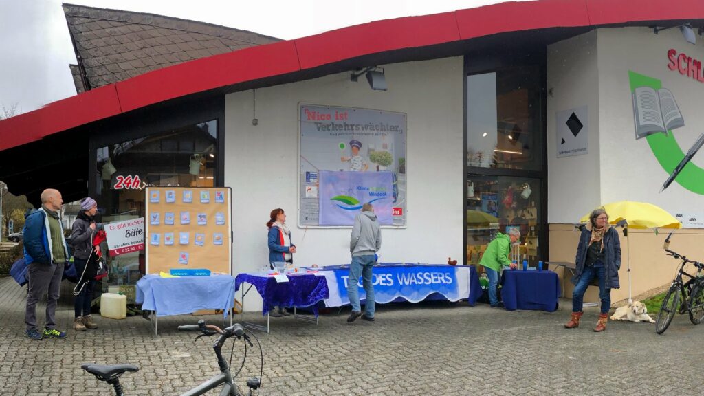 Unser Infostand vor der Buchhandlung Schlösser bestehend aus einer Reihe von Tischen mit verschiedenen Informationsmaterial und Experimenten bzw. Quizzes. Vor dem Infostand stehen einige Menschen.