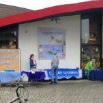 Unser Infostand vor der Buchhandlung Schlösser bestehend aus einer Reihe von Tischen mit verschiedenen Informationsmaterial und Experimenten bzw. Quizzes. Vor dem Infostand stehen einige Menschen.