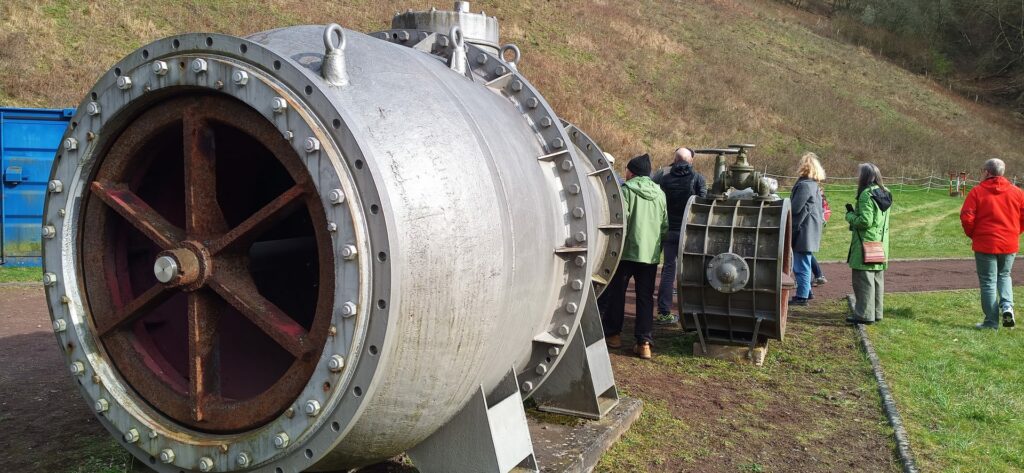 Ein metallisches Bauteil (die Turbine) im Vordergrund und mehrere Menschen im Hintergrund, die stehend in der Turbine Platz hätten.