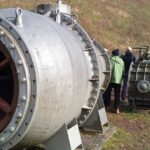 Ein metallisches Bauteil (die Turbine) im Vordergrund und mehrere Menschen im Hintergrund, die stehend in der Turbine Platz hätten.