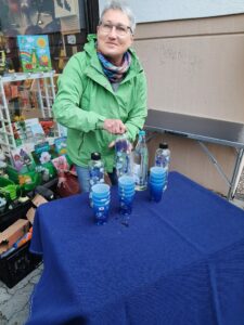 Eine Frau in grüner Jacke steht vor einem Tisch mit blauer Tischdecke. Auf dem Tisch stehen mehrere Flaschen mit Wasser gefüllt und mehrere Becher.